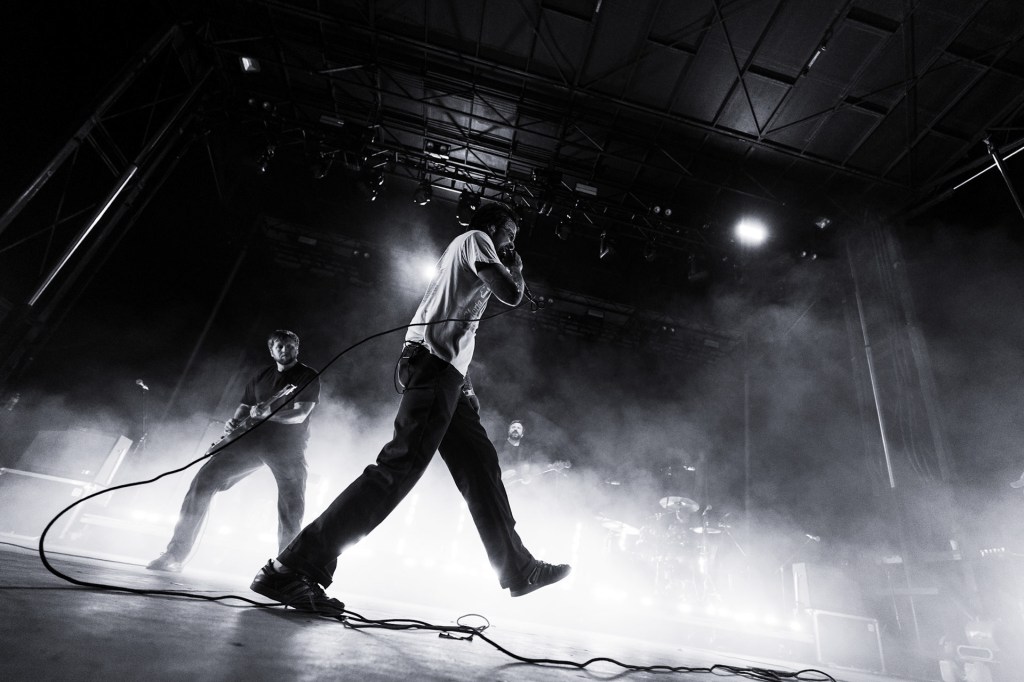 A black and white image of a live band performing on stage, with one vocalist in the foreground and a guitarist slightly behind. The stage is filled with smoke and bright stage lights, creating an energetic atmosphere.