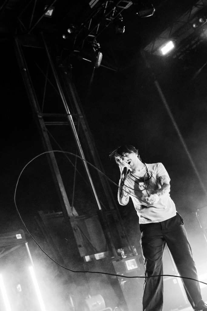 A black and white image of a singer performing on stage, holding a microphone and wearing a casual outfit. The background features stage lights and equipment, creating a dynamic atmosphere.