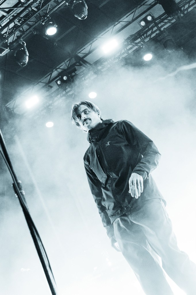 A male performer stands on stage in a foggy, dimly lit environment, with lights directing down. They are wearing a dark jacket and look intently at the audience.