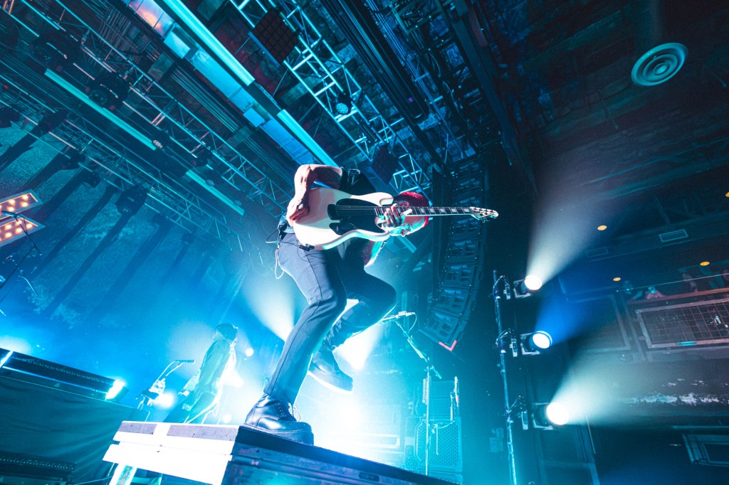 A guitarist performing energetically on stage under vibrant blue lighting, mid-jump with a white electric guitar.