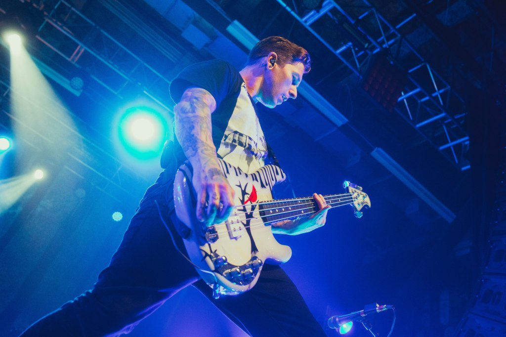 A male musician playing a white bass guitar on stage with dramatic blue lighting and a lively atmosphere.