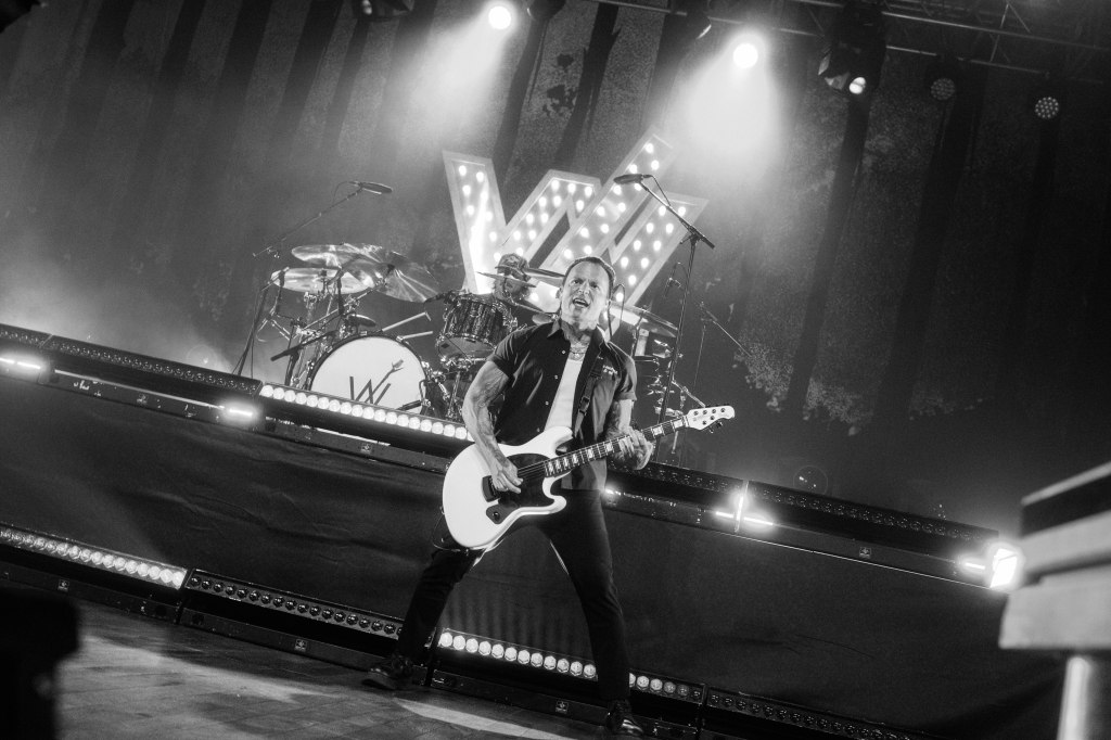 A guitarist performing on stage with a white electric guitar, in front of a drum set, under dramatic lighting. The background features a large illuminated letter 'W'.