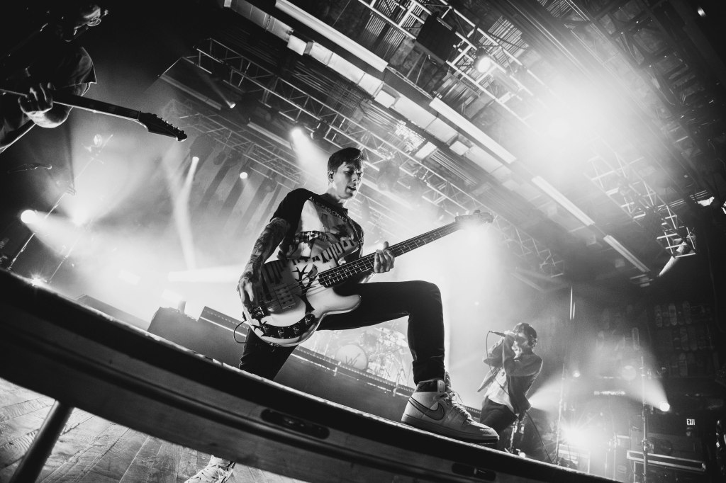 A bassist performing on stage during a concert, with a lead vocalist in the background, illuminated by dramatic stage lights in a black and white setting.