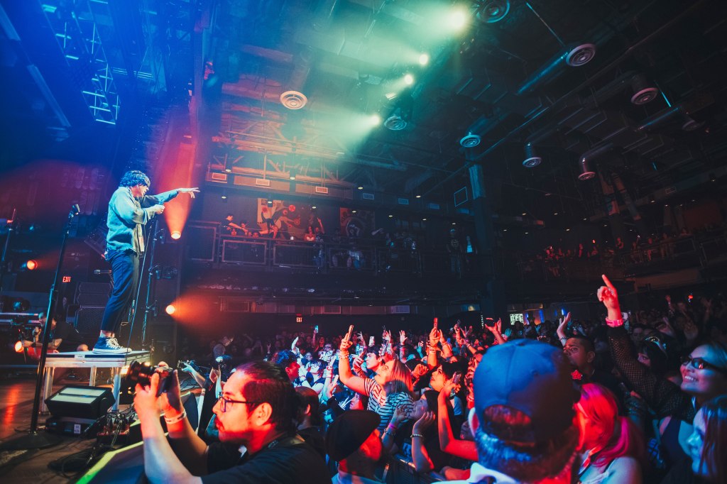 A musician performing on stage with a microphone in hand, while fans in the audience cheer and raise their hands. Colorful stage lights illuminate the scene, creating an energetic atmosphere.