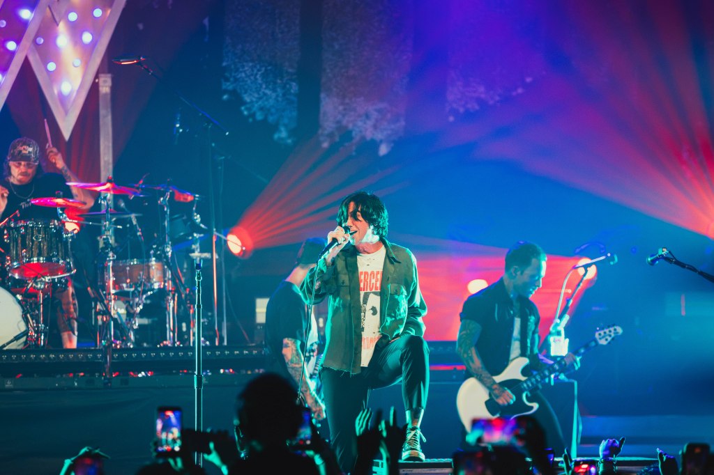 A rock band performs on stage with vibrant lighting and energetic crowd; the lead singer engages with the audience while the drummer plays in the background.