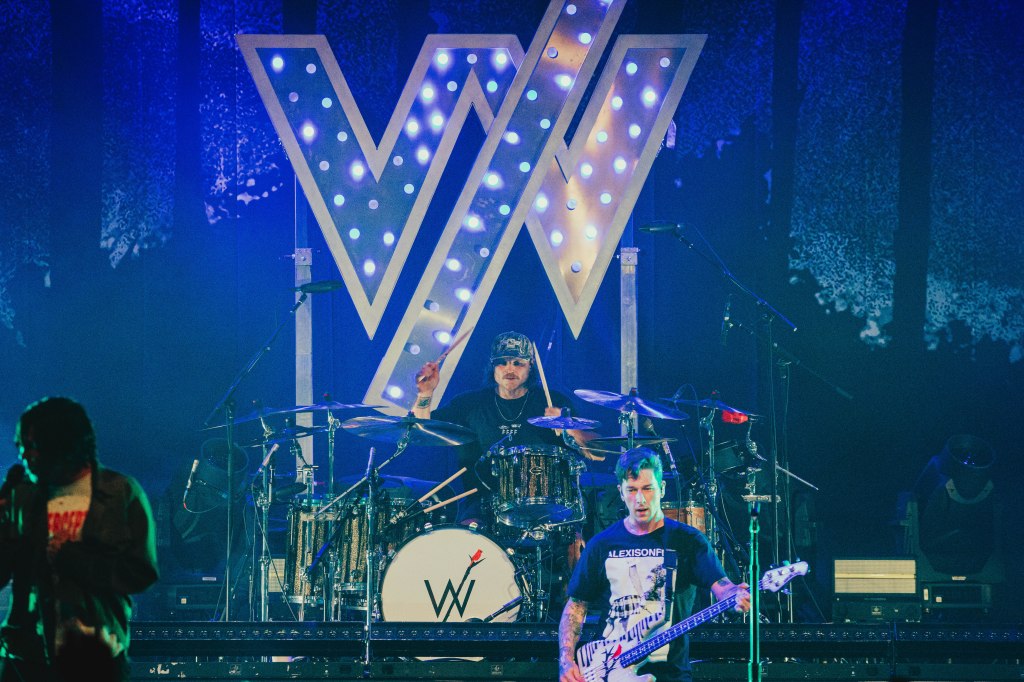 A band performing live on stage with a drummer and a bassist, featuring a large illuminated letter 'W' backdrop.