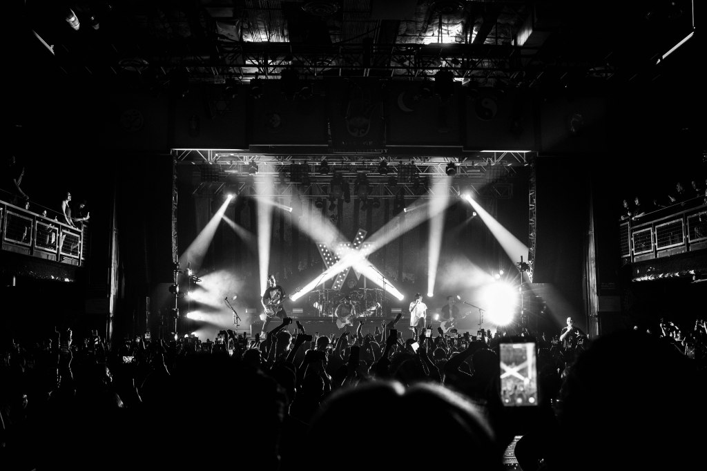 A black and white image of a live concert, featuring a band performing on stage with bright stage lights and a crowd of fans raising their hands in excitement.