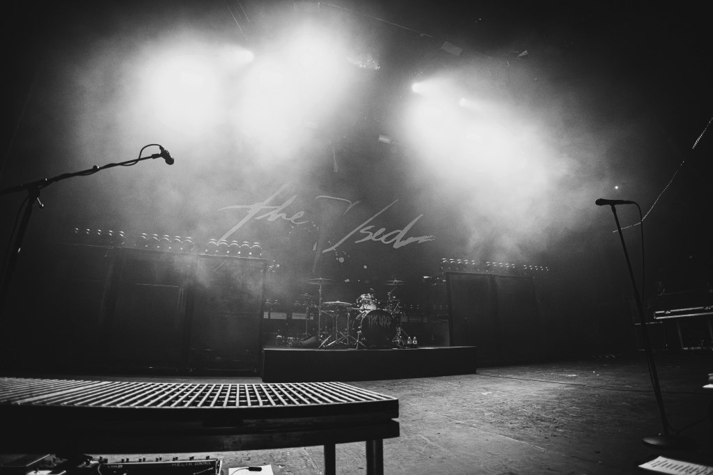 A concert stage set up with a microphone stand, drum kit, and amplifiers, surrounded by stage lights and fog, giving a moody atmosphere.