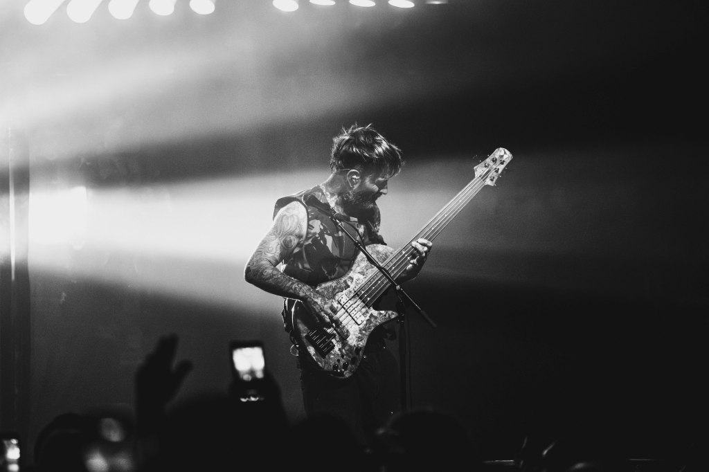 A musician playing a bass guitar on stage, highlighted by dramatic lighting, in a black and white photography style.