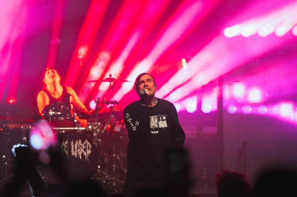 A live music performance by a band featuring a vocalist singing passionately on stage, with a drummer in the background. Colorful stage lights create a vibrant atmosphere.
