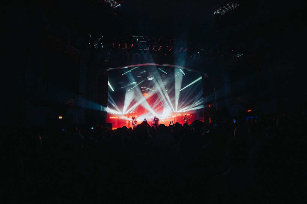A live music performance on stage with bright colored lights and a crowd of fans in the foreground.