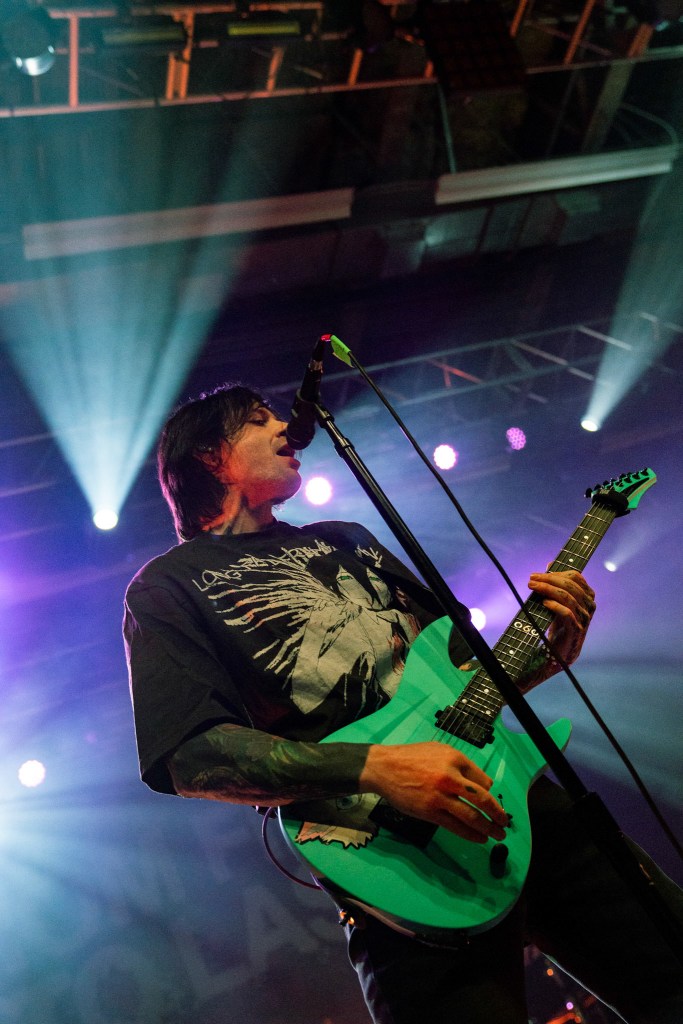 Musician performing on stage with an electric guitar, dressed in a band t-shirt, singing into a microphone under colorful stage lights.
