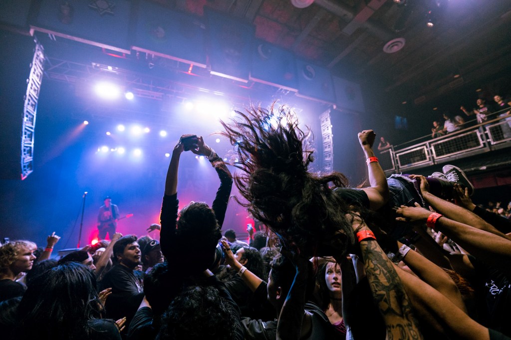 A crowd at a concert, with a person crowd surfing overhead and others reaching up, illuminated by stage lights.