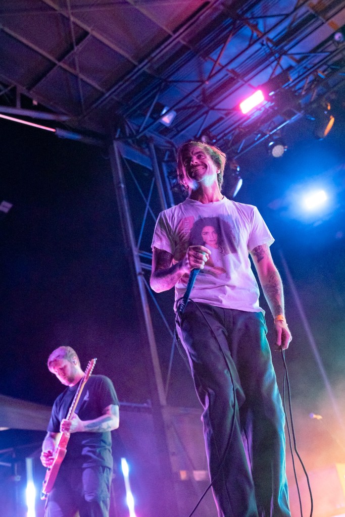 Lead singer performing on stage with a microphone, wearing a graphic tee, while a guitarist plays in the background under colorful stage lights.