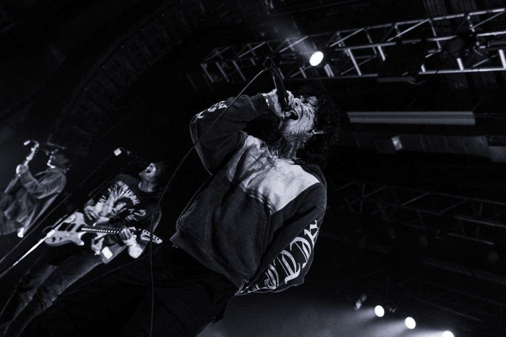 A vocalist performing passionately on stage, surrounded by band members with guitar and drums, set against dramatic stage lighting.