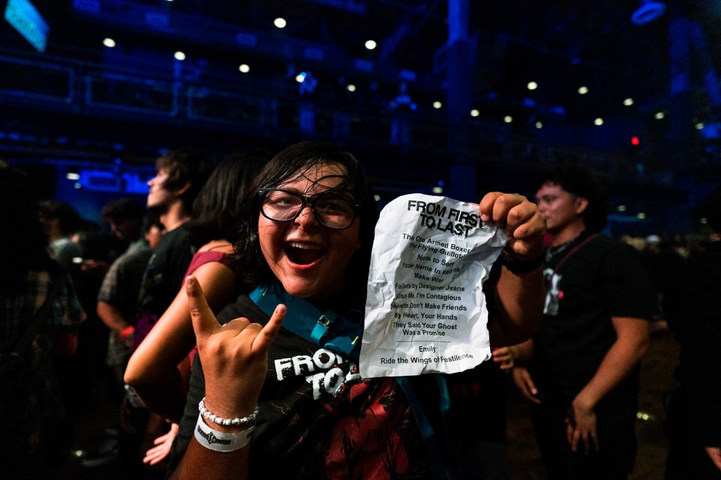 A cheerful fan at a concert holds up a setlist from the band 'From First to Last,' making a rock hand gesture, with a crowd visible in the background.