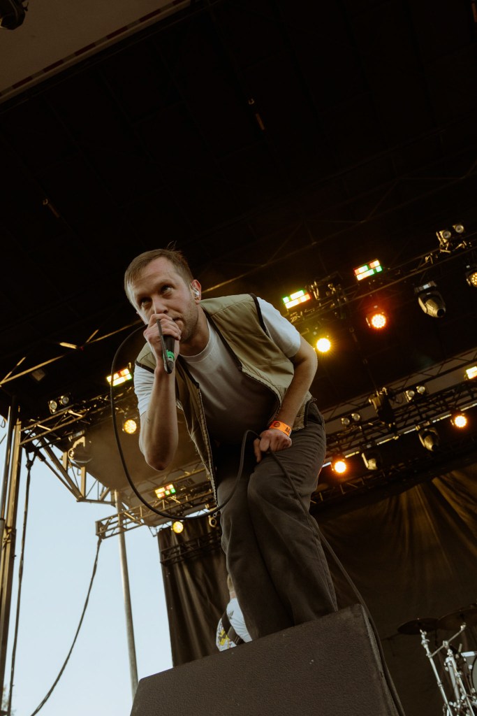 A singer performing on stage with a microphone, wearing a vest and pants, with colorful stage lights in the background.