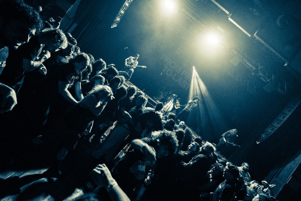 A crowded concert scene with energetic crowd members dancing in front of a stage, illuminated by bright stage lights.