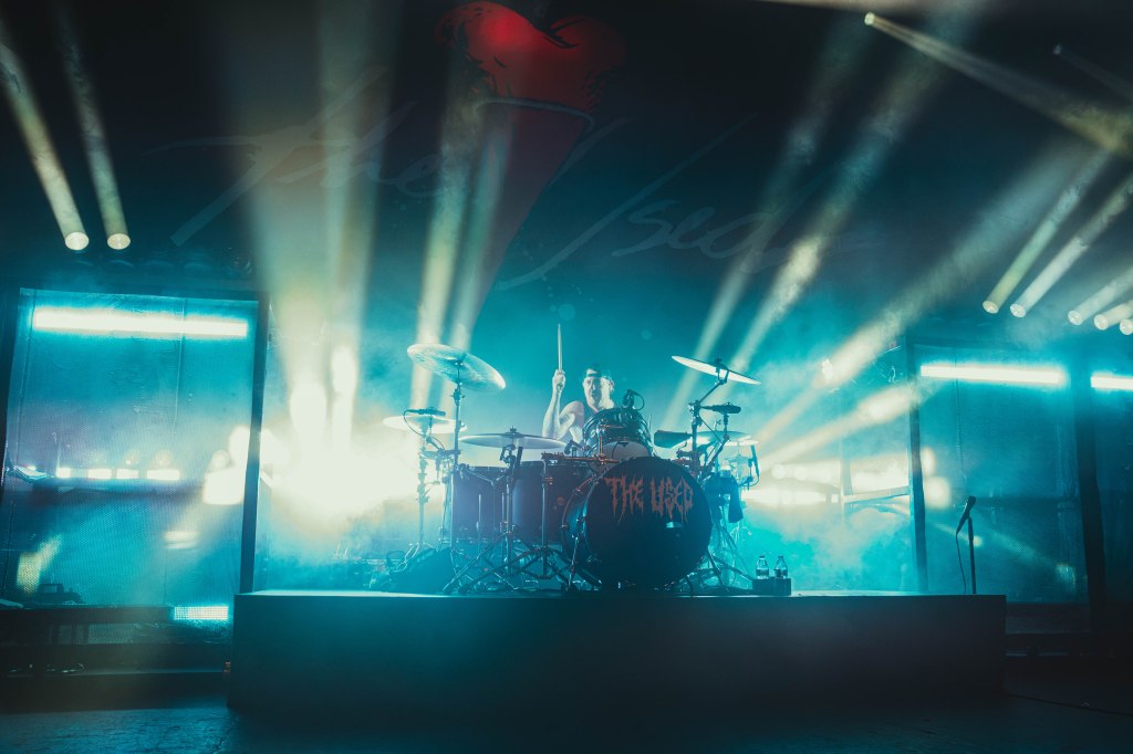 A drummer performs on stage surrounded by vibrant lights and smoke during a concert.