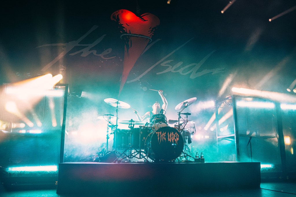 A drummer performing on stage with dramatic lighting and smoke, in front of a backdrop featuring 'The Used' logo.