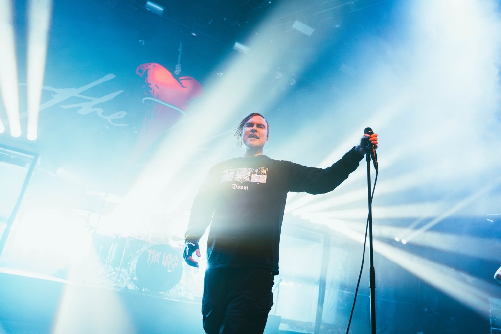 A vocalist performing on stage, holding a microphone and wearing a black long-sleeve shirt with graphic designs, surrounded by dramatic lighting and fog.