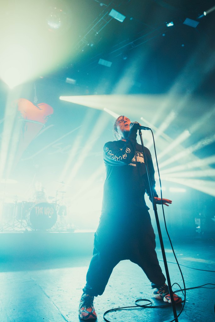 A male singer passionately performing on stage, holding a microphone with dramatic lighting and visual effects in the background.