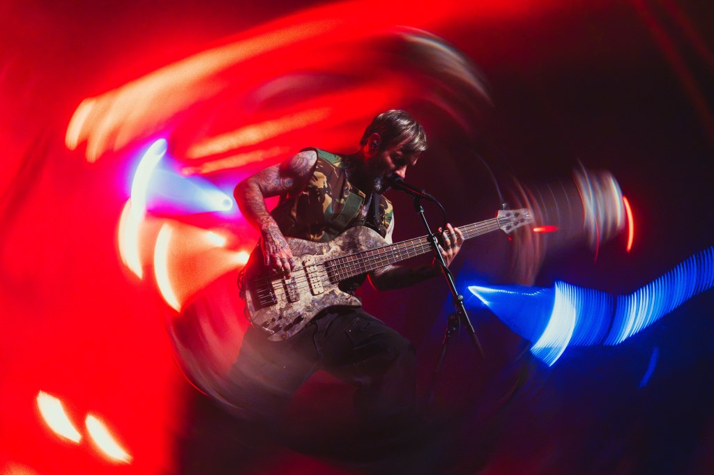 A musician playing an electric bass guitar on stage, surrounded by colorful lights with a blurred motion effect.