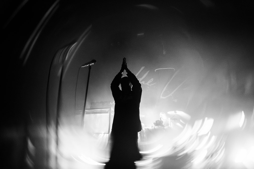 Silhouette of a performer with hands raised, standing on stage amidst swirling smoke and lighting effects during a concert.