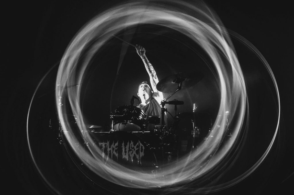 A drummer passionately performing on stage, surrounded by a swirling light effect in black and white.