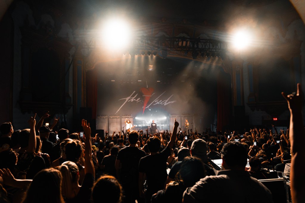 A large crowd enjoys a live concert in a theater, with a performer on stage illuminated by bright lights and the audience raising their hands.