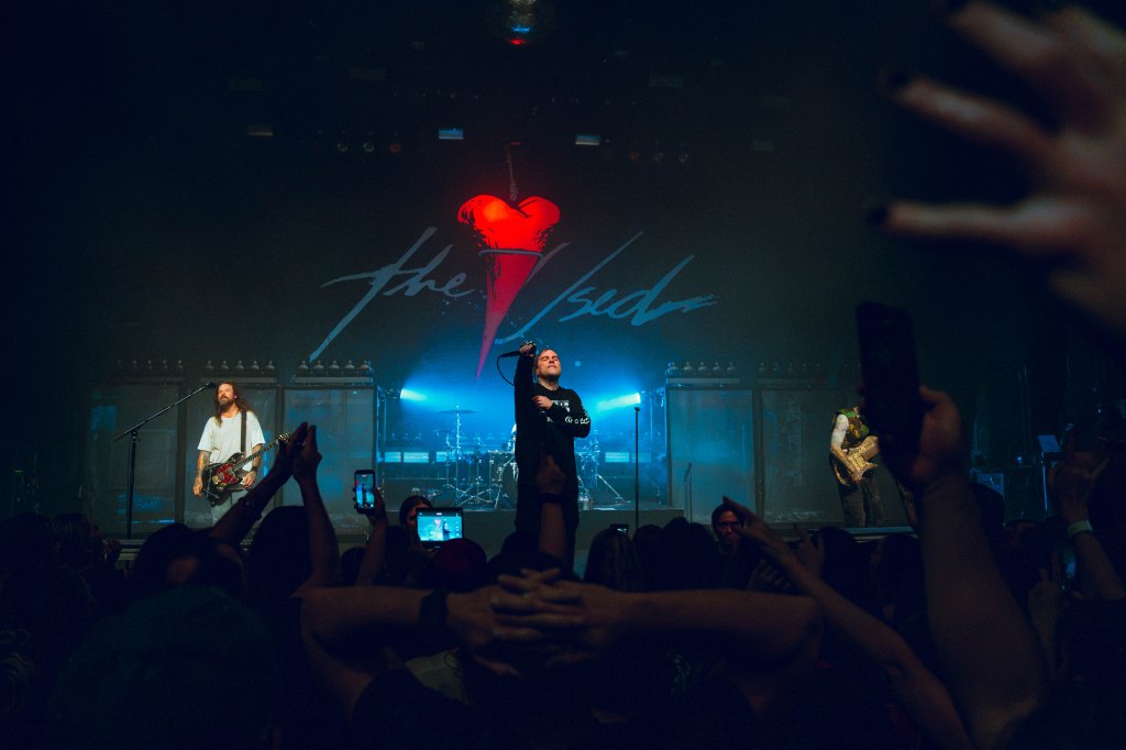 A rock band performing on stage with dramatic lighting, featuring a lead singer and two musicians, while audience members hold up their phones to capture the moment.