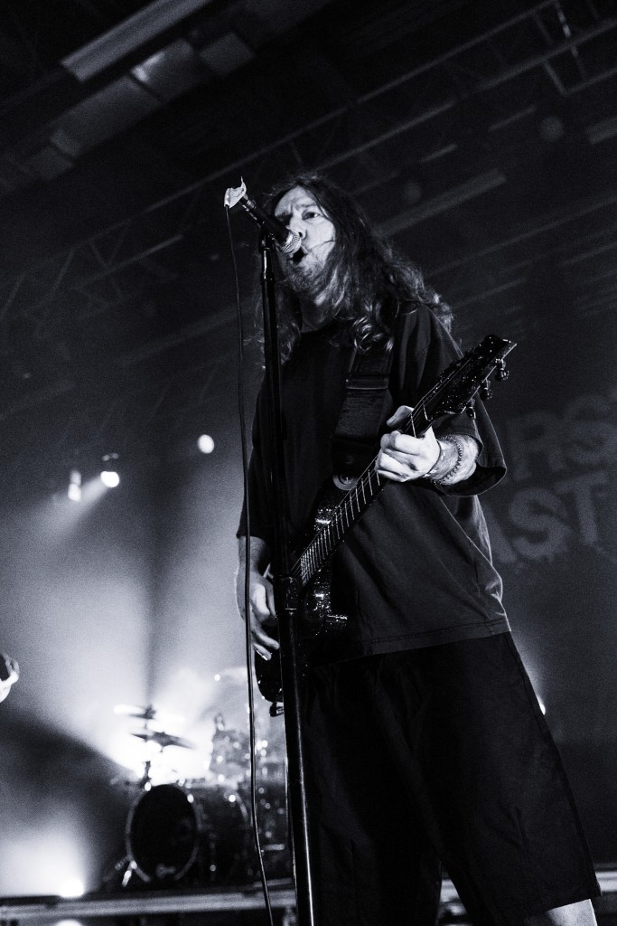 A musician performing on stage with long hair, holding a guitar and singing into a microphone, illuminated by stage lights in a dark setting.
