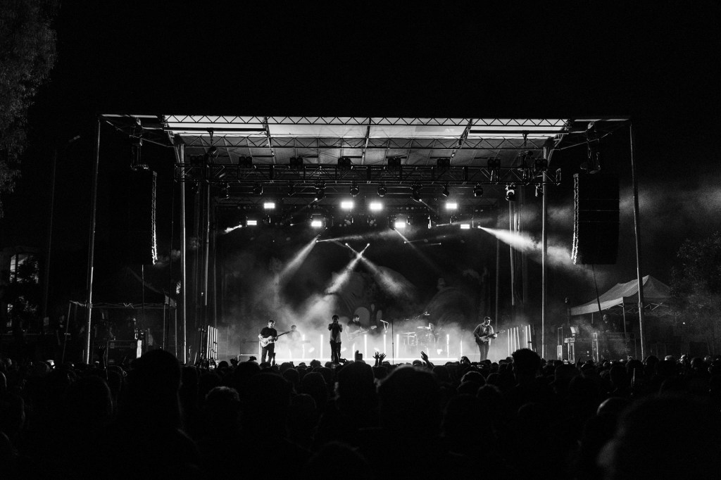 Black and white image of a live music concert at night, featuring a band on stage with bright spotlights and an audience captured in front.