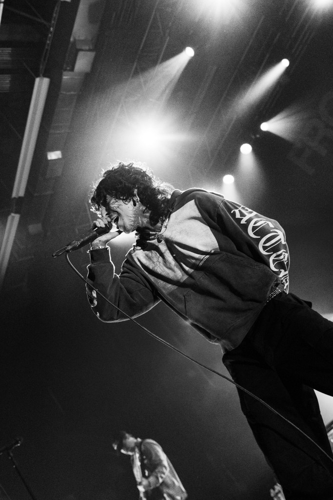 A singer performing on stage with a microphone, wearing a hoodie, surrounded by dramatic lighting.