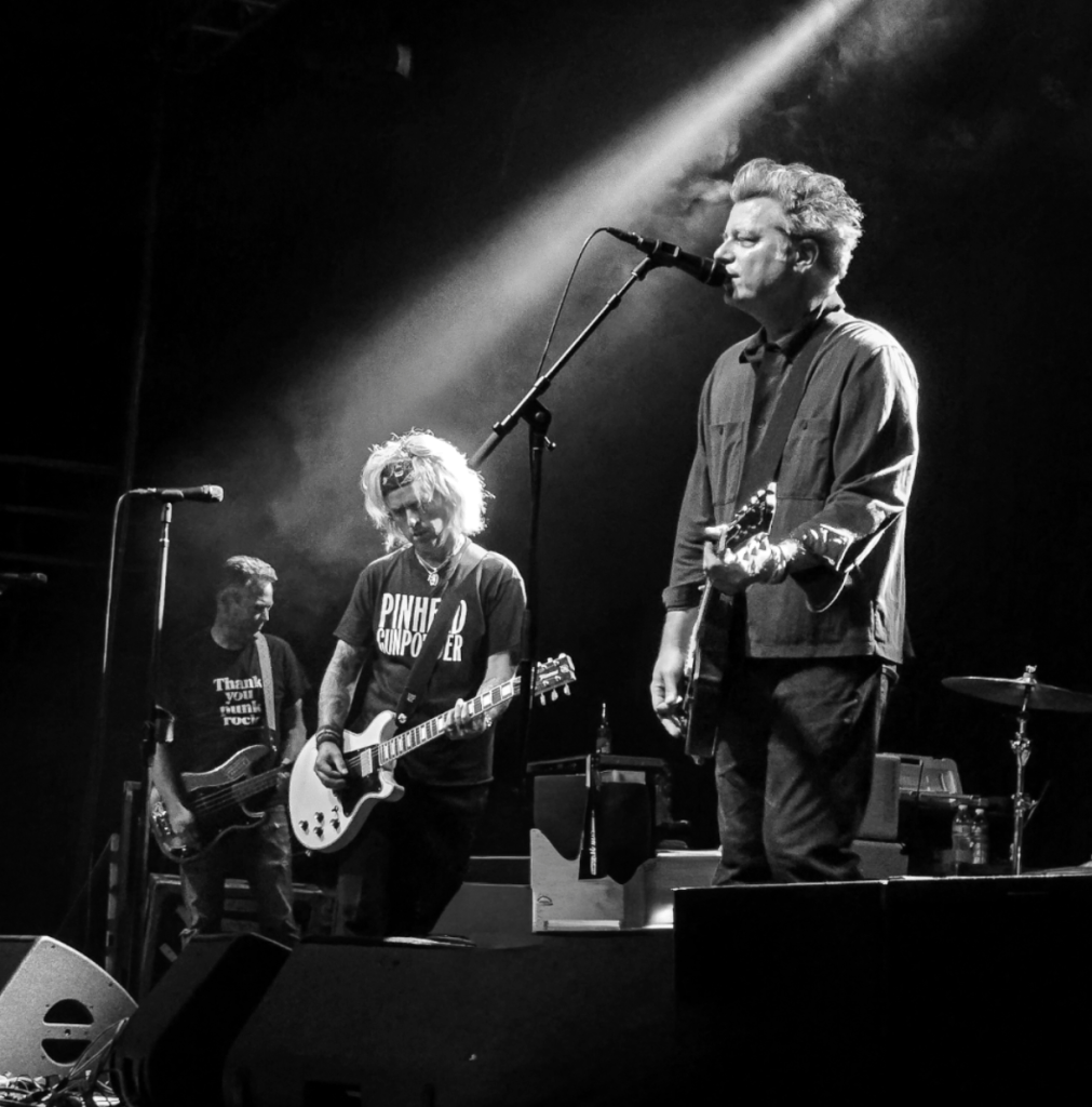 Black and white image of a rock band performing on stage. The lead singer stands at the microphone while two guitarists play beside him. The bassist is visible in the background, and stage lighting creates a dramatic effect.